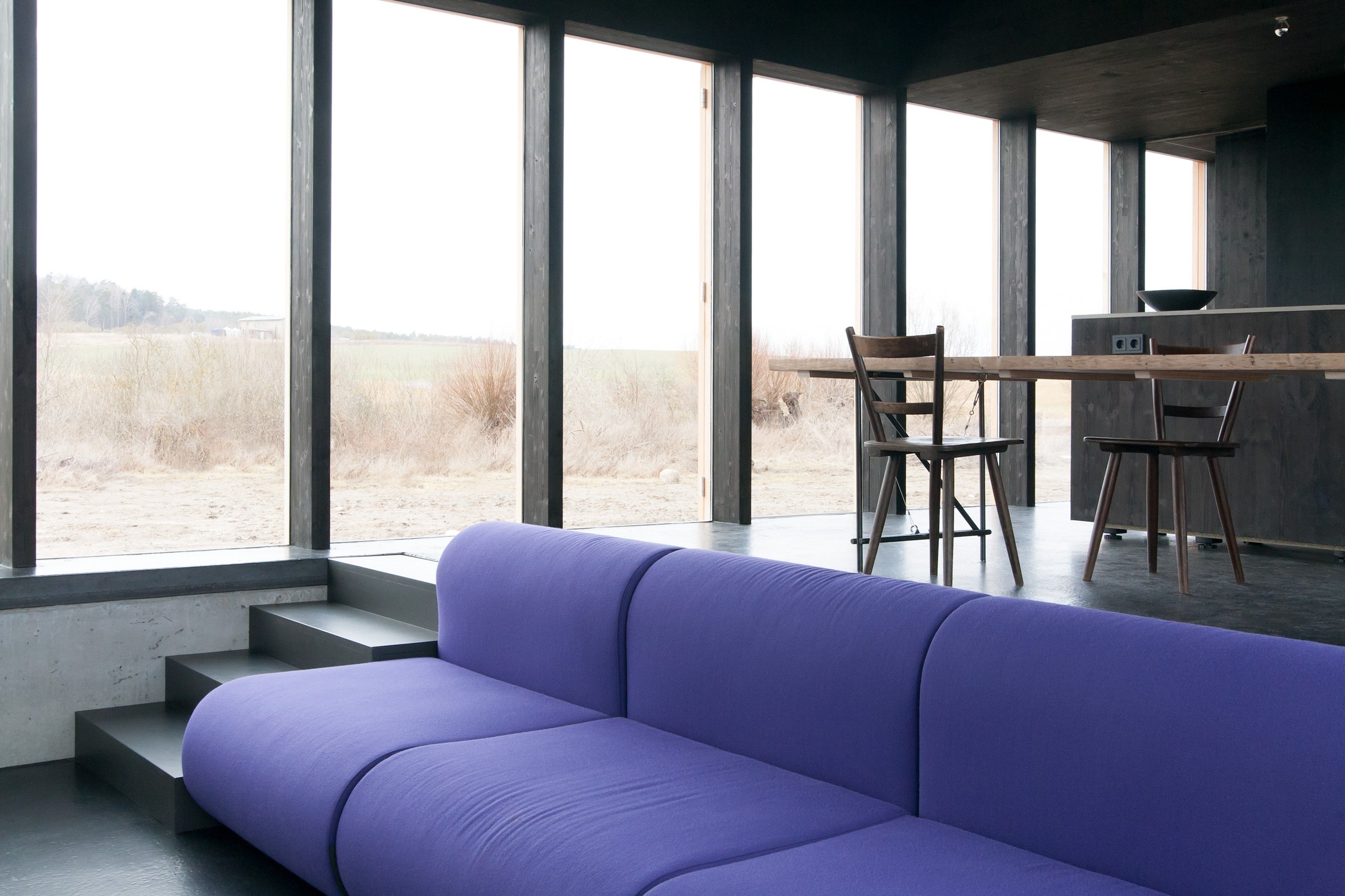 Ein modernes Interieur mit einem violetten Sofa, einem Holzesstisch mit Stühlen und großen Fenstern mit Blick auf ein Feld.