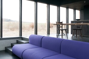 Ein modernes Interieur mit einem violetten Sofa, einem Holzesstisch mit Stühlen und großen Fenstern mit Blick auf ein Feld.
