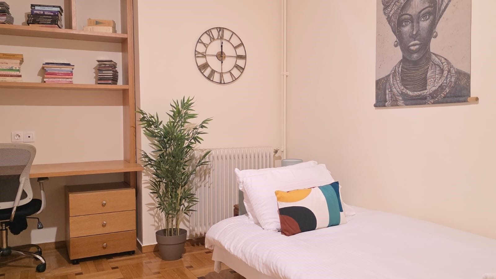 A bedroom with a bed, a desk, a chair, a potted plant, a wall clock, and a painting.
