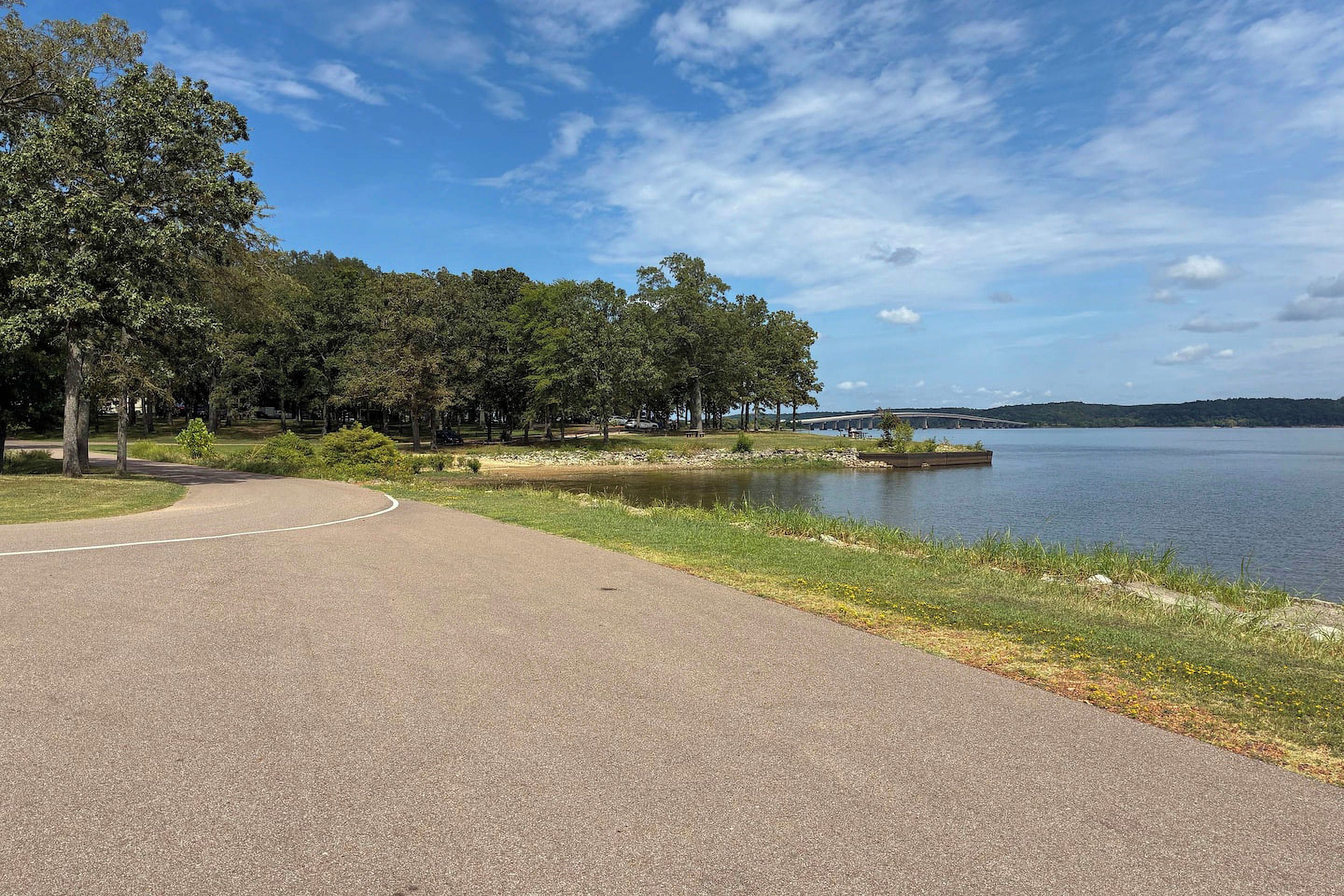 A paved pathway alongside a body of water with trees and a distant bridge.