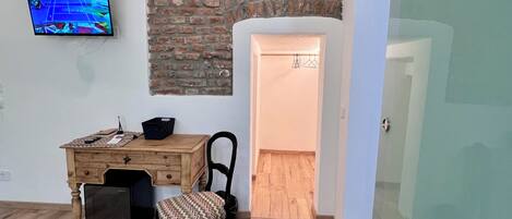 A room with a wooden desk, a chair, a television mounted on the wall, and a brick wall.