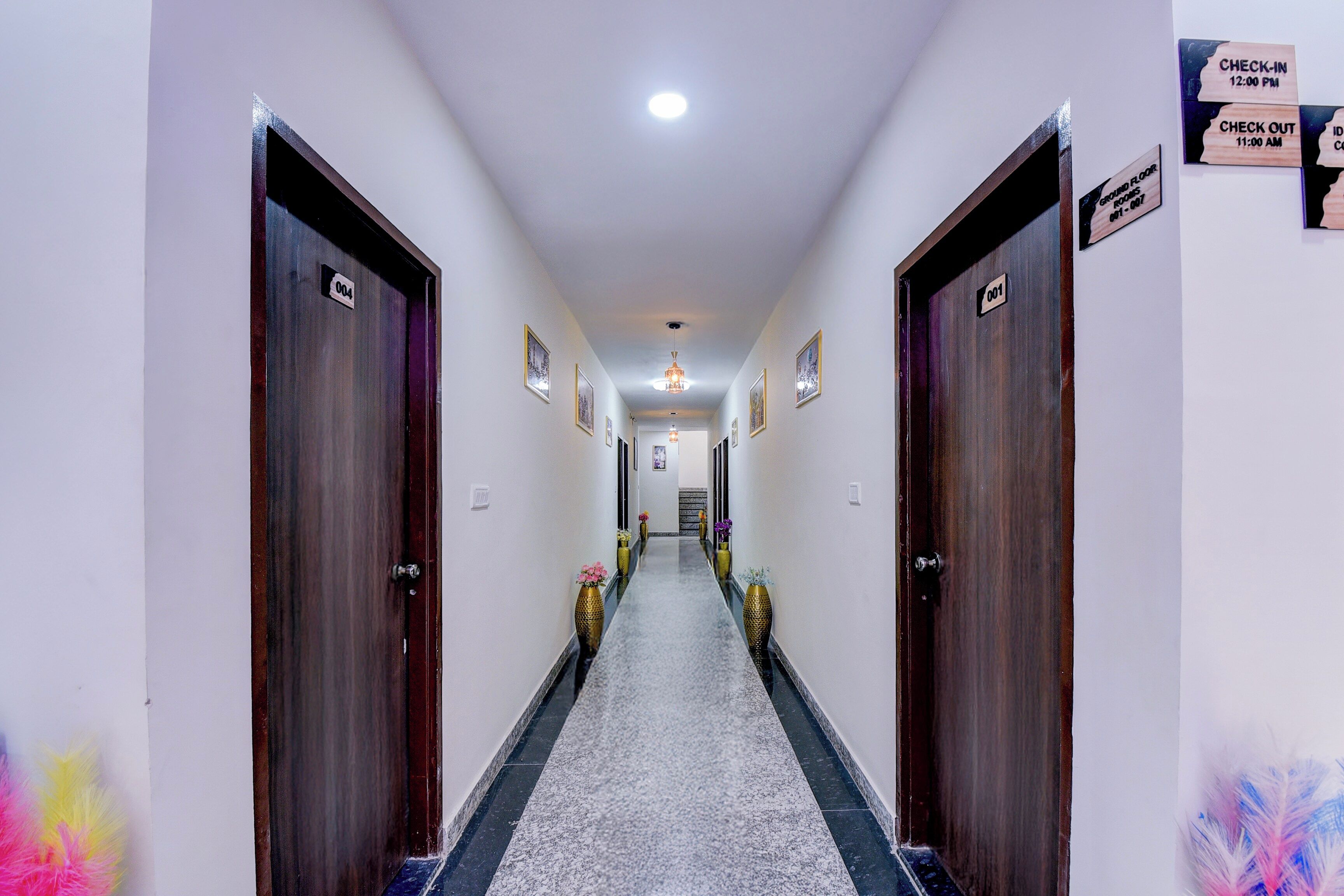 A hotel hallway with doors numbered 001 to 004, check-in and check-out signs, and framed pictures on the walls.