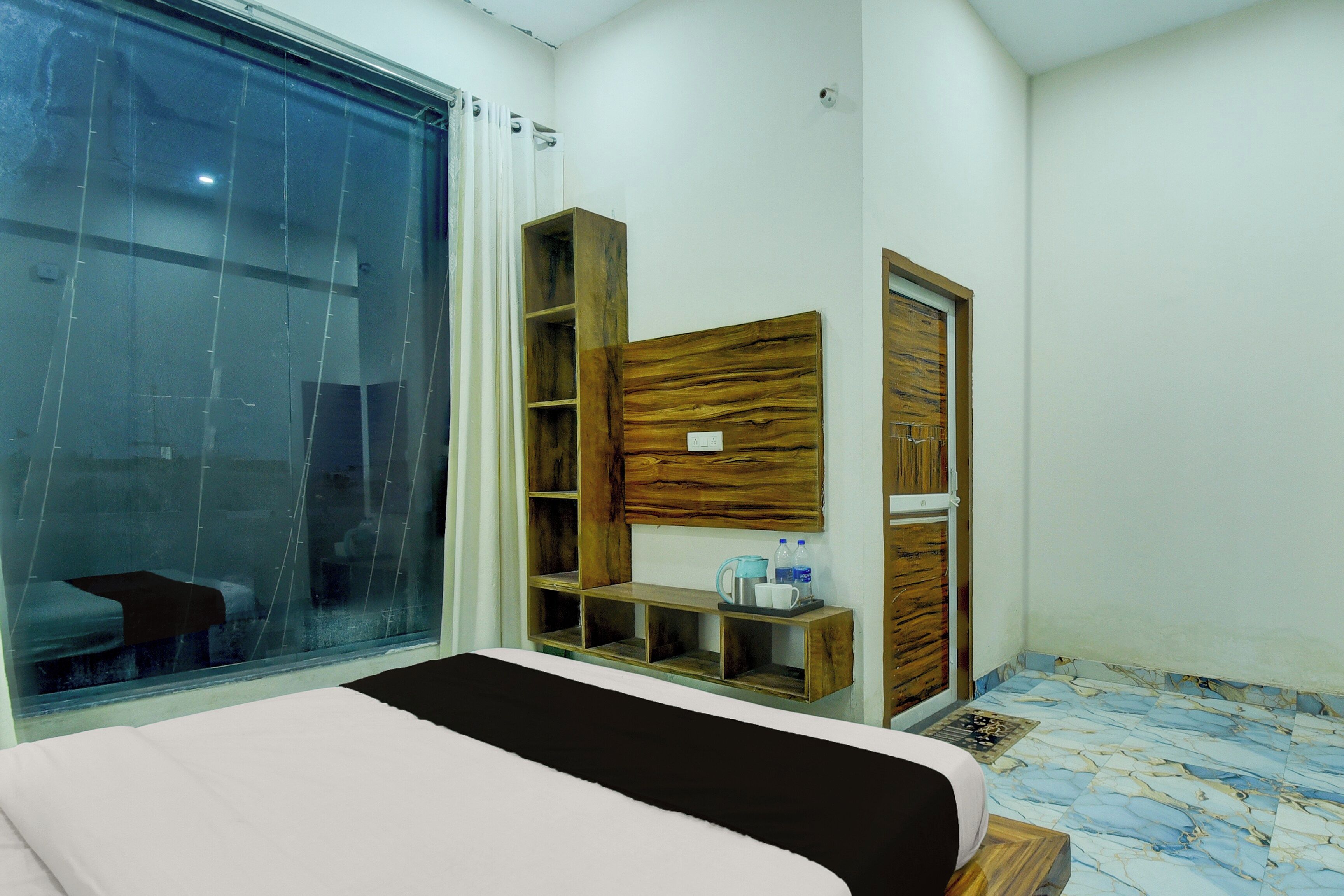 Habitación de hotel con cama, armario de madera, estante con artículos de aseo y un ventanal grande.