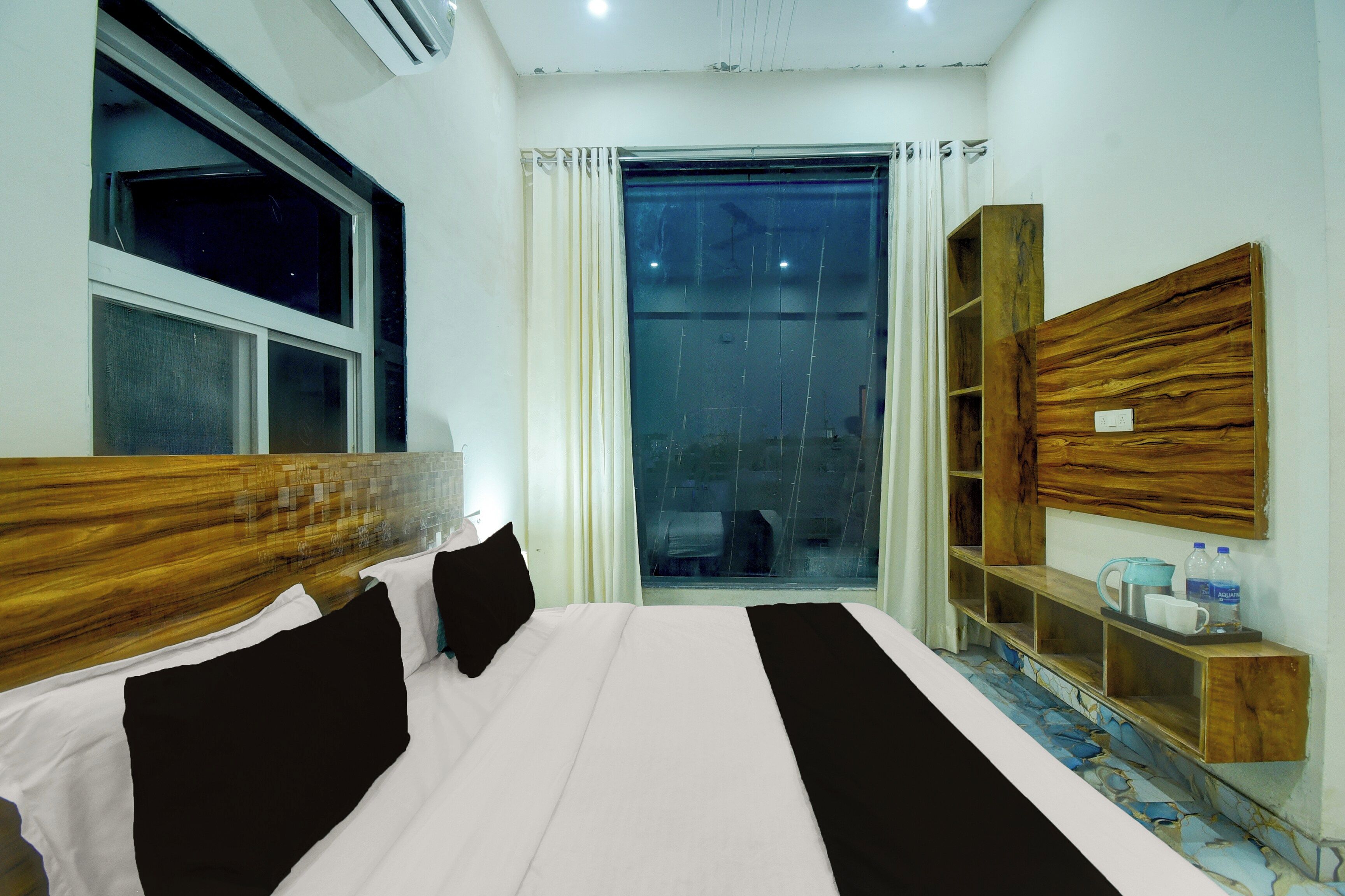 Habitación de hotel con una cama grande, cabecera de madera, una ventana con cortinas, un estante de madera con objetos y vistas al exterior.