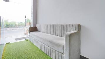 A corner of a room with a grey upholstered bench against a white wall, a green artificial grass area, and a window with a view of a street and buildings.