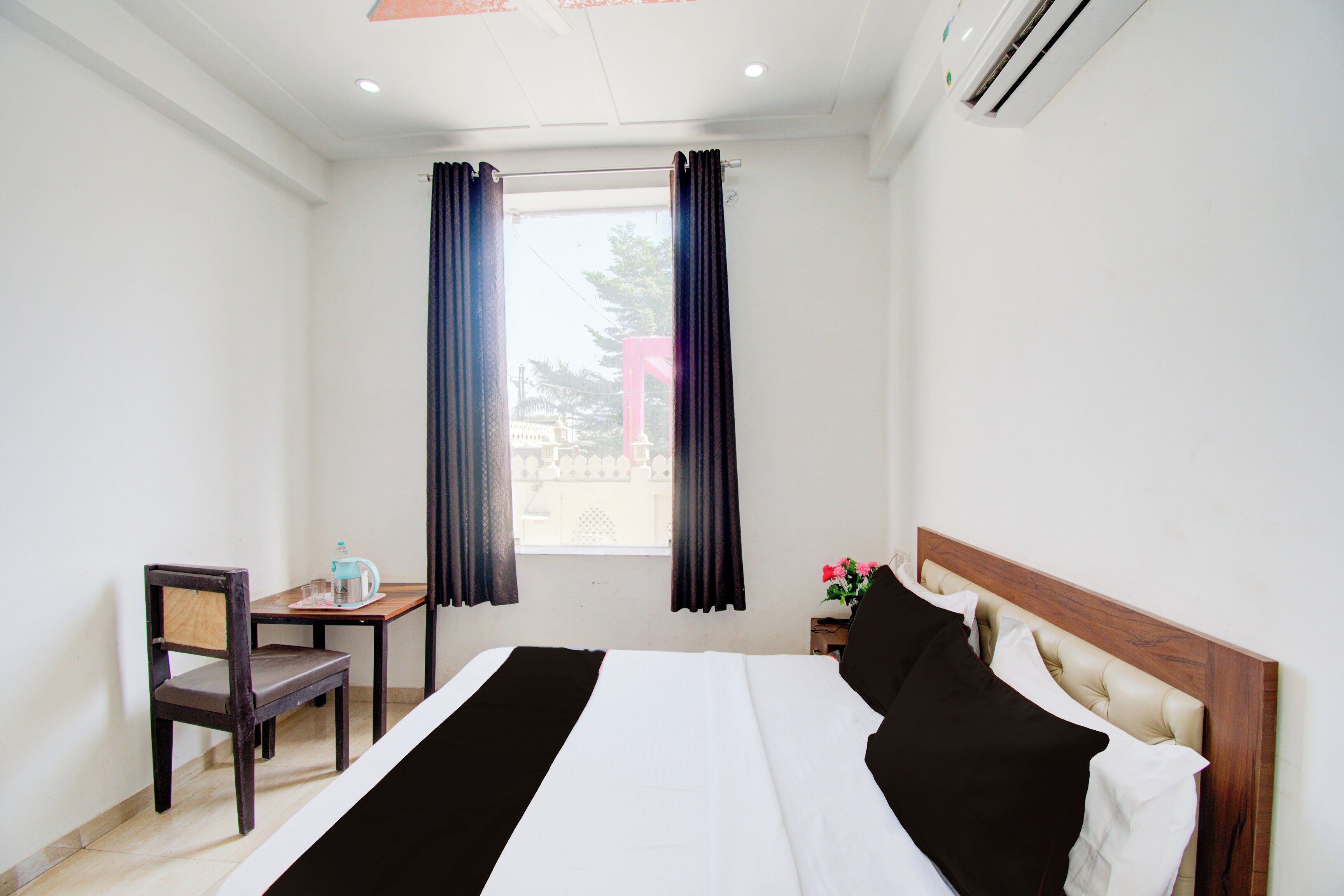 A hotel room with a bed, a chair, a small table with a kettle, and a window with curtains.