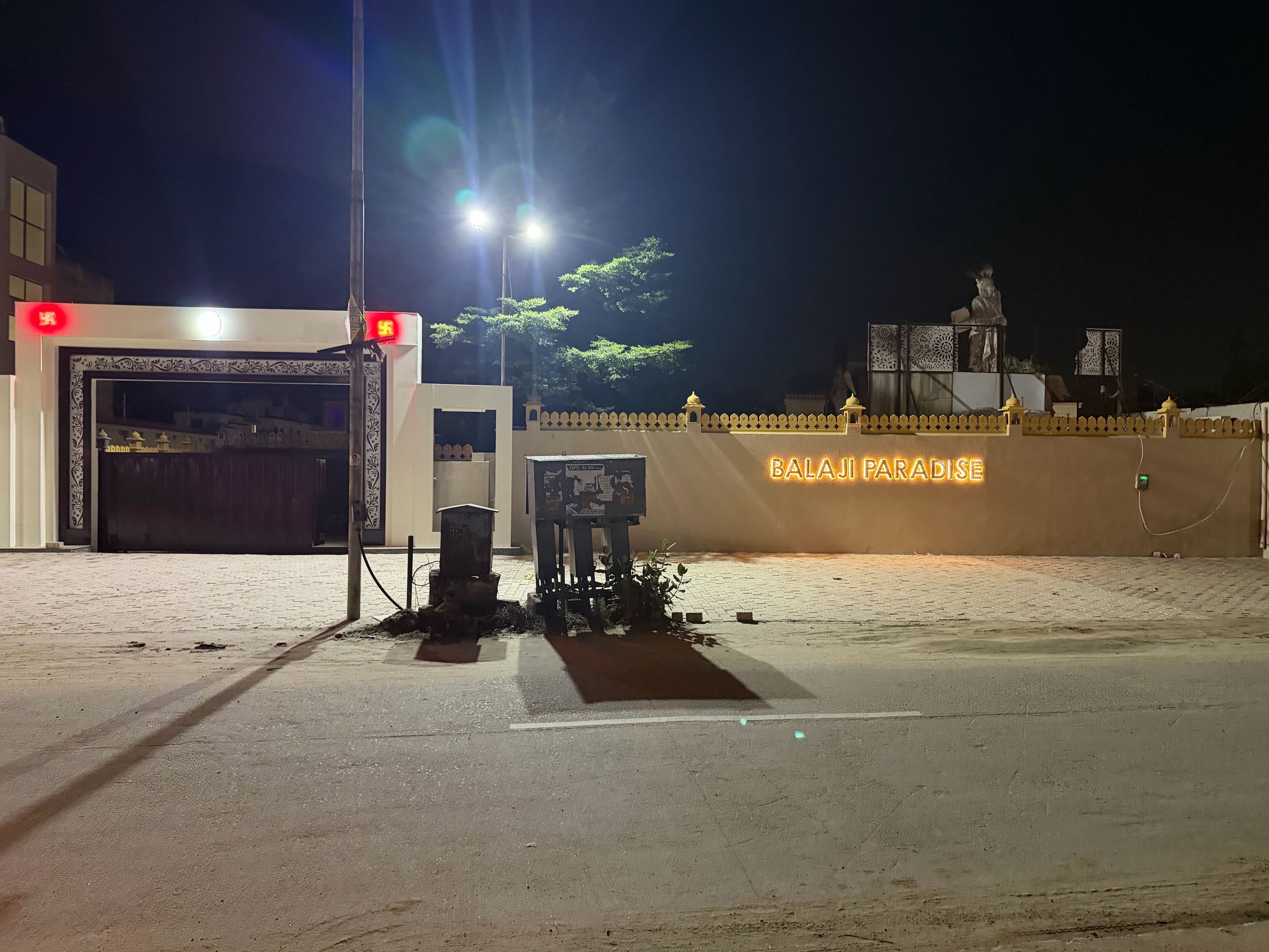 A gated entrance with a neon sign for 'BALAJI PARADISE' and a statue in the background.