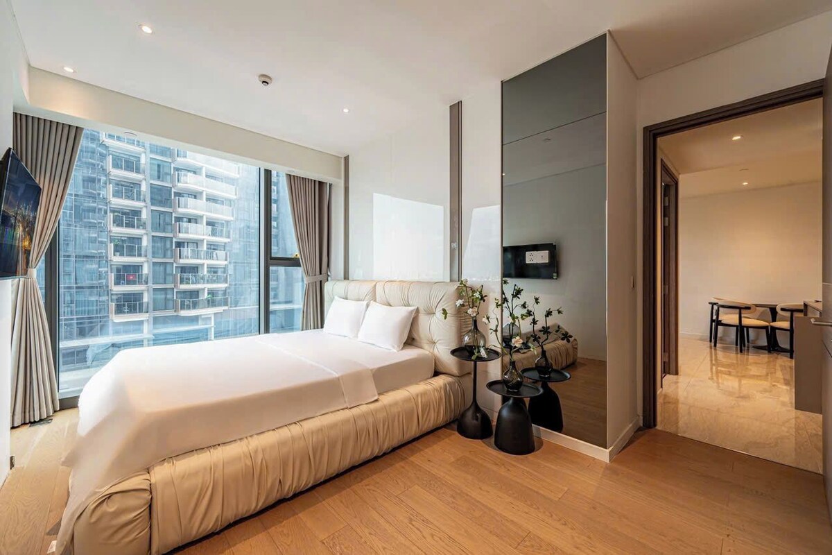 A modern hotel room with a large bed, a dining area, and a view of a high-rise building.