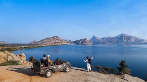 A jeep parked on a rocky shore overlooking a calm lake with mountains in the distance. - Bookmark Resorts Cheetahgarh Jawai (Bali)