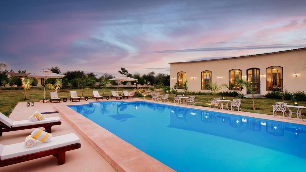 Piscina con sillones de descanso y sombrillas, rodeada de un jardín ajardinado y un edificio con ventanas arqueadas.