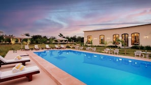 A swimming pool with lounge chairs and umbrellas, surrounded by a landscaped garden and a building with arched windows. - Bookmark Resorts Cheetahgarh Jawai (Bali)