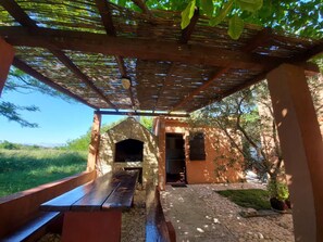 Un patio al aire libre con un banco de piedra y un horno de ladrillo, rodeado de follaje y un cielo despejado.