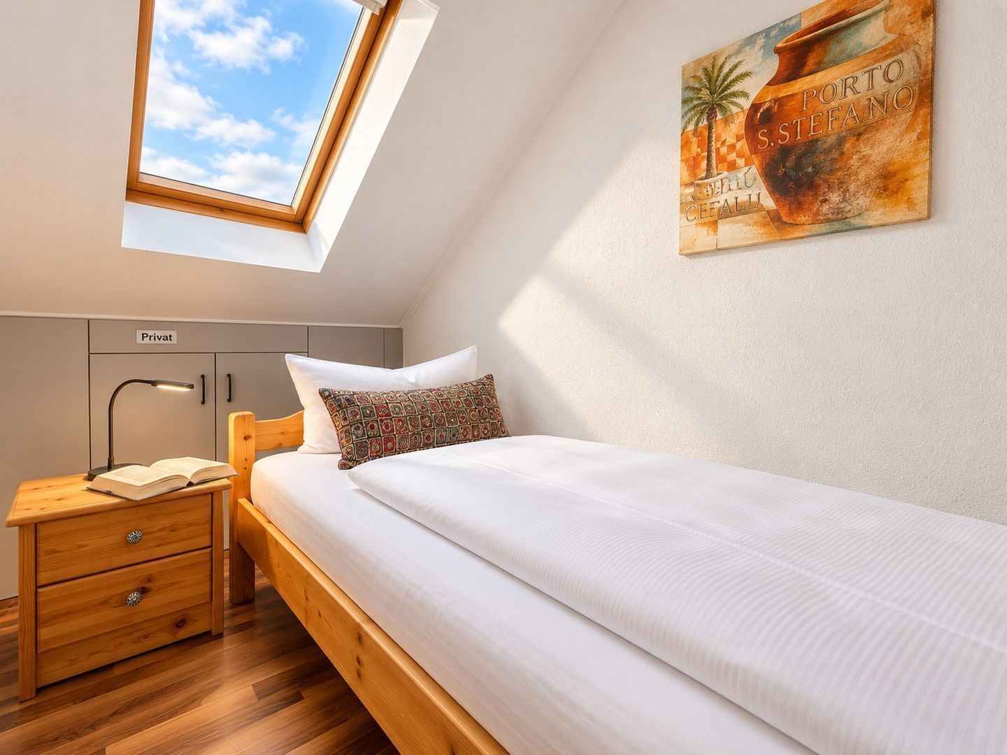 Ein Schlafzimmer mit Dachfenster, einem Bett mit weißer Bettwäsche, einem Nachttisch mit Lampe, einer Holzkassette und einem Wandregal.