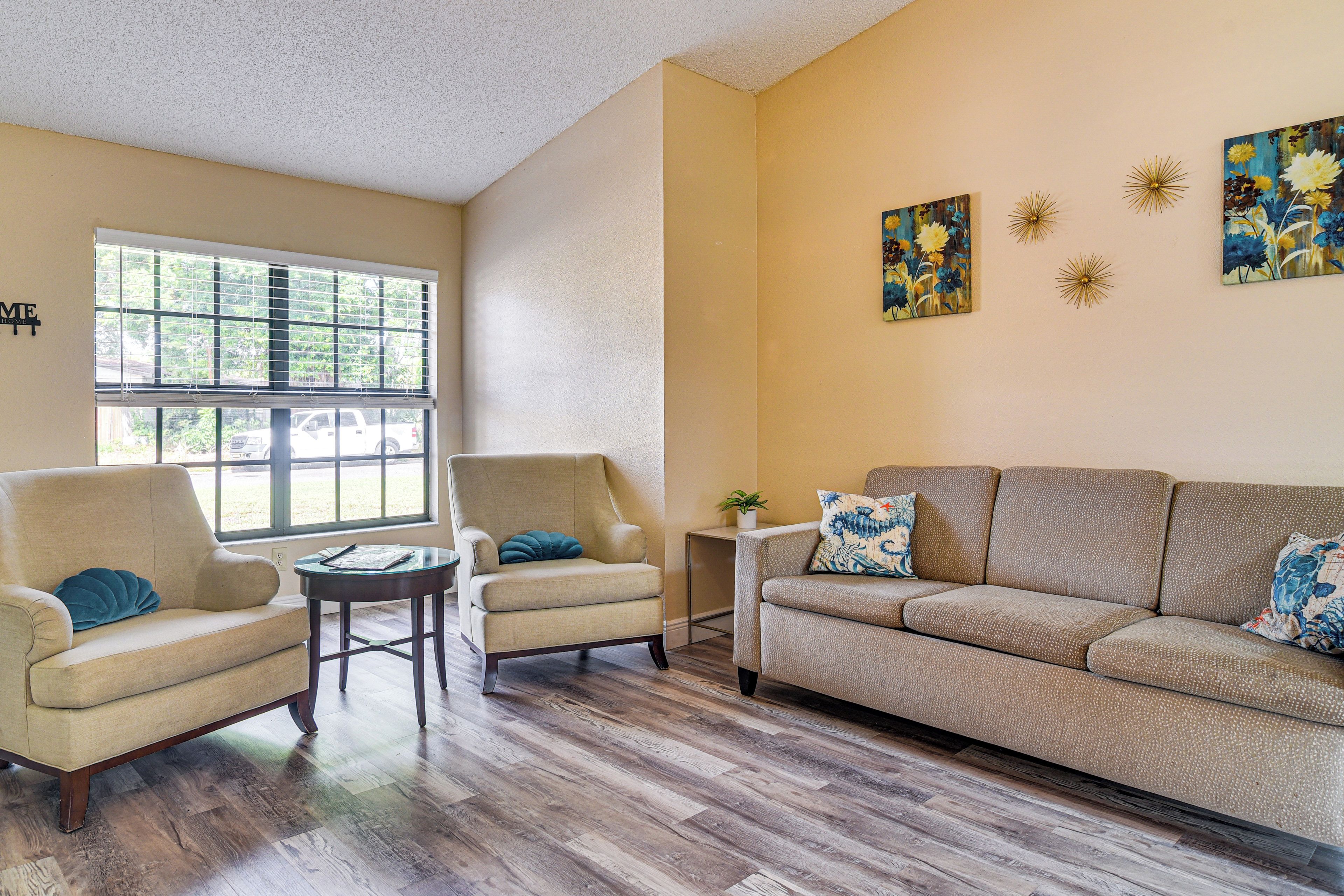 A living room with a beige sofa, two armchairs, a coffee table, and a small side table with a plant. There are large windows with blinds, a ceiling with a textured finish, and wall-mounted decorations.