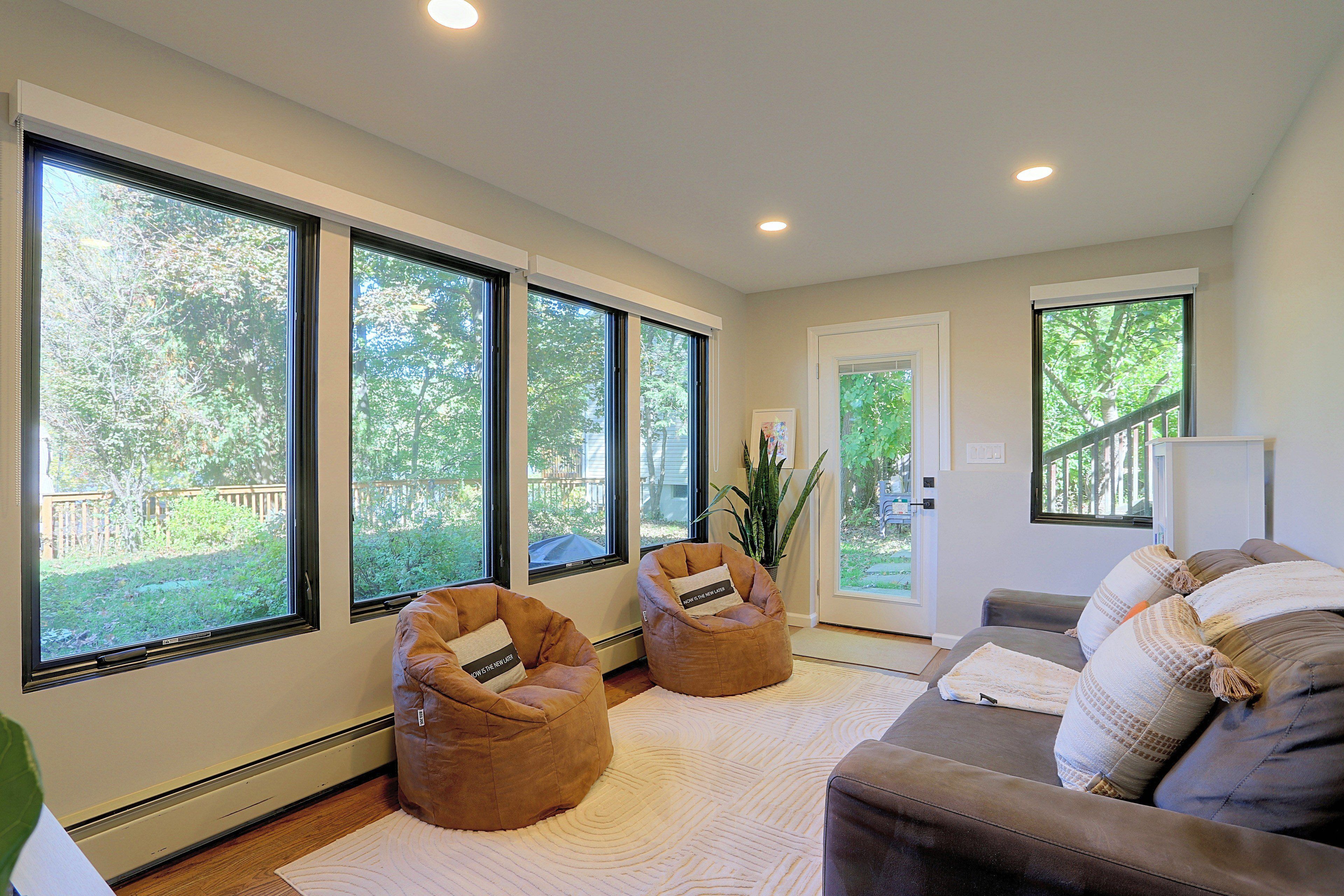 A spacious living room with large windows, a grey sofa, two bean bags, and a potted plant.