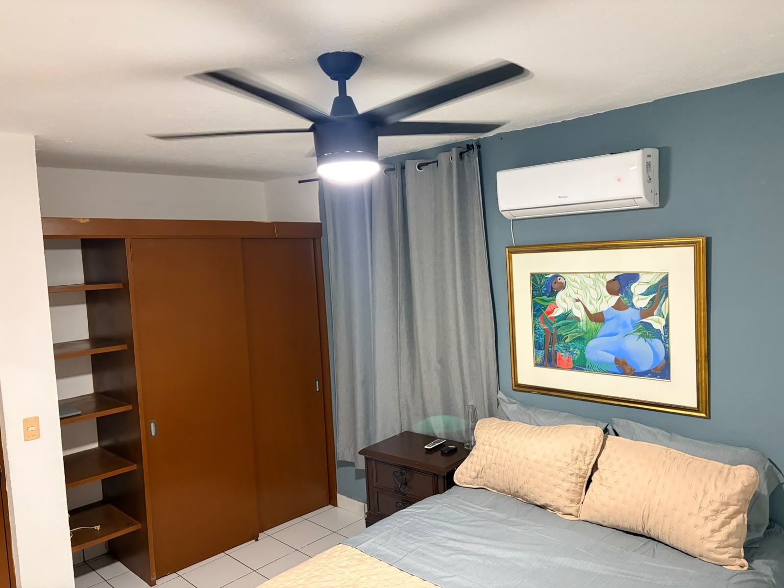 A bedroom with a bed, a wardrobe, a ceiling fan, a framed picture, and an air conditioning unit.