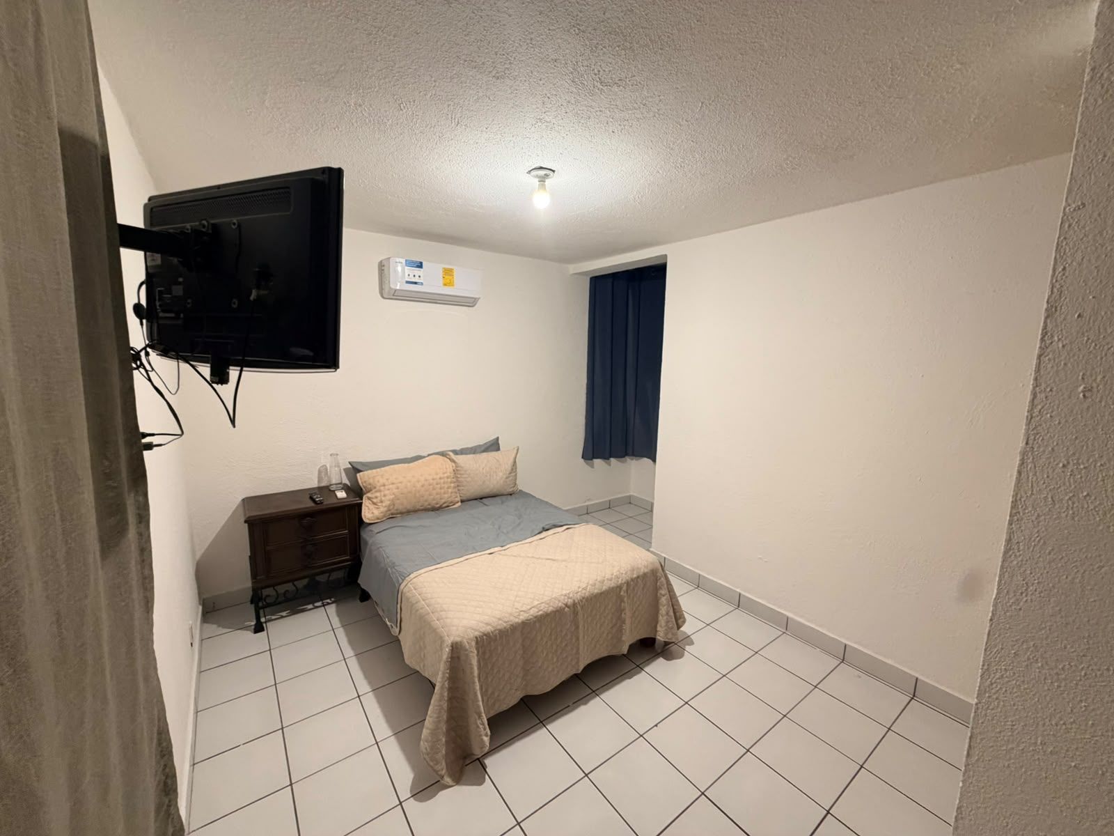 A bedroom with a bed, a bedside table, a TV mounted on the wall, and a ceiling light.