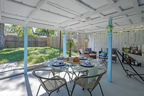 Ein überdachter Patio-Bereich mit Esstisch, umgeben von Korbstühlen und einer Sitzecke mit Kissen und einem kleinen Tisch.