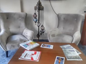 Una habitació amb dues cadires tapissades de color gris, una taula de fusta amb papers i llibres, i una làmpada decorativa penjant.