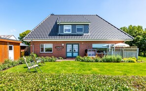 Ein zweigeschossiges Haus mit einem Ziegeldach, einer Haustür und einem kleinen Garten mit einer Bank und Blumentöpfen.