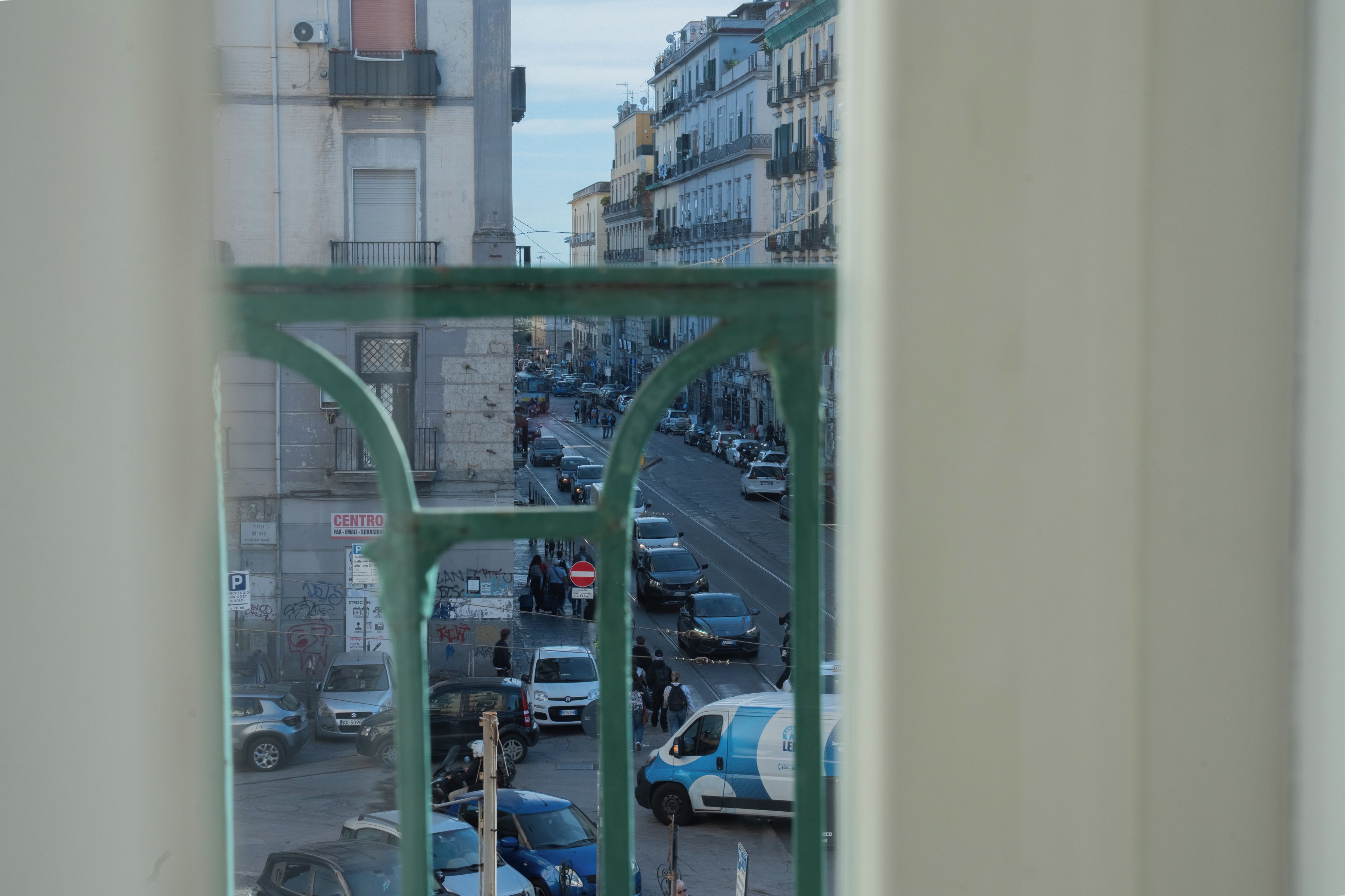 A street view through a window with a metal frame, featuring parked cars, a blue van, and buildings with balconies.