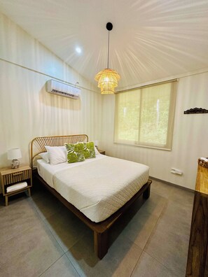 Une chambre à coucher comprenant un lit, une table de chevet, une lampe, un appareil de climatisation, une fenêtre avec des rideaux et un luminaire suspendu.