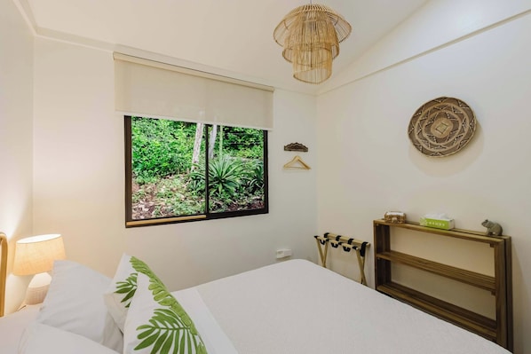 Quarto com cama, mesa de cabeceira, luminária pendente, janela com vista para vegetação e um prato decorativo fixado na parede.