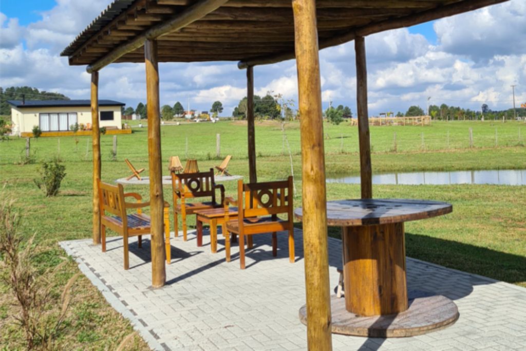 Un pabellón de madera al aire libre con una mesa y sillas, rodeado de césped y una masa de agua.
