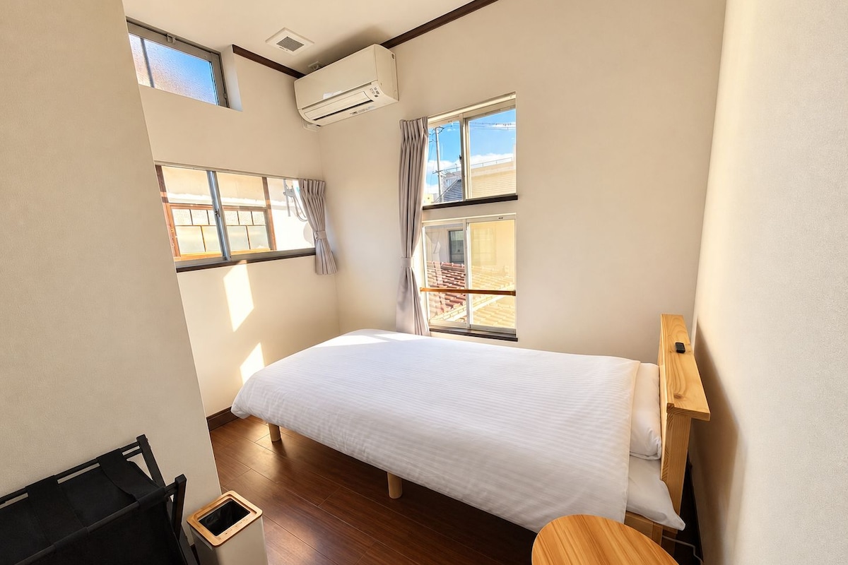 A bedroom with a bed, a window, a small table, and a trash bin.