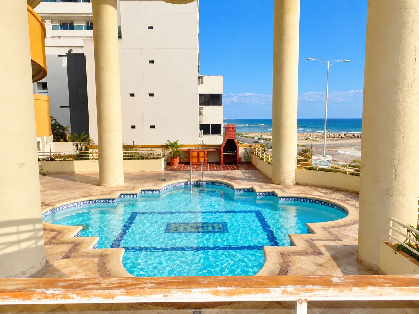 A swimming pool with a tiled deck, a barbecue area, and a view of the beach and ocean.