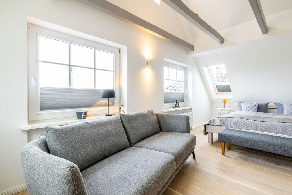 Une chambre moderne avec un canapé gris, un lit avec une couette grise et une fenêtre donnant sur un immeuble.