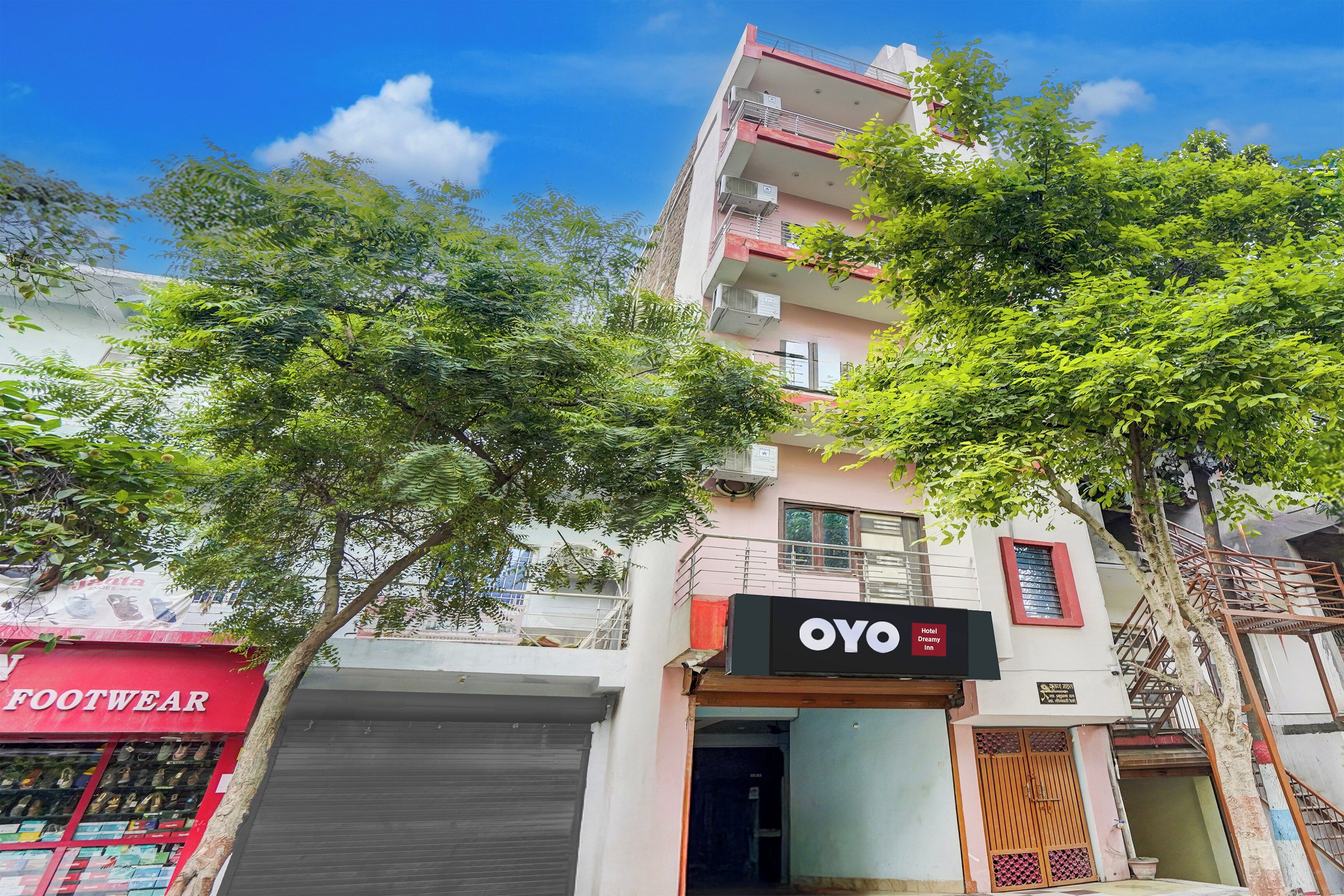 Un hotel llamado OYO con un letrero, una fachada roja con la leyenda 'FOOTWEAR' a la izquierda y un árbol en primer plano.
