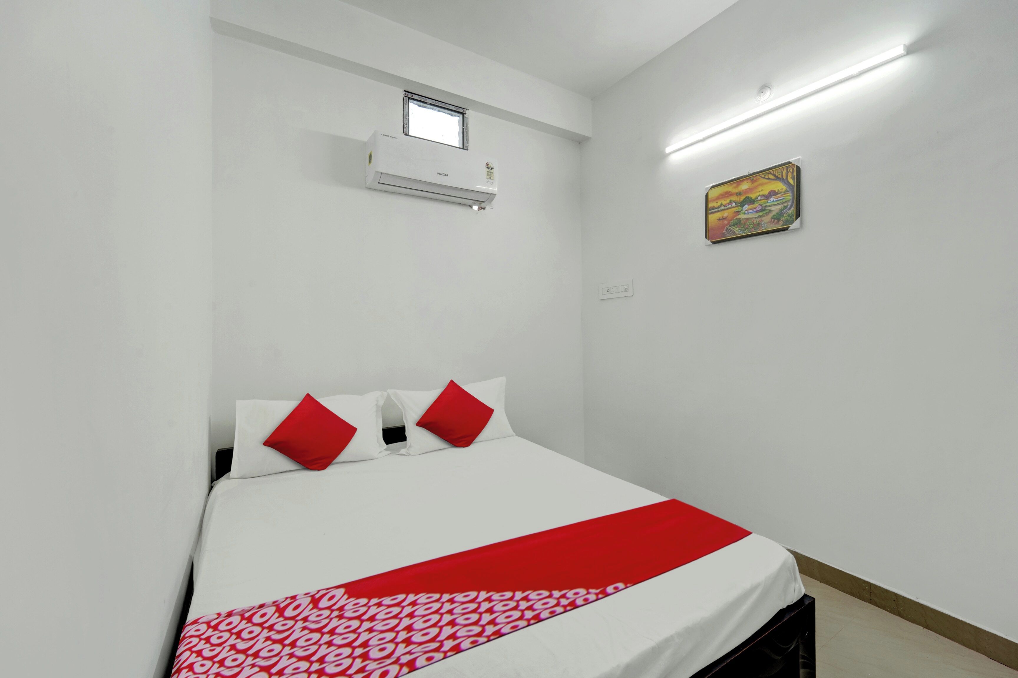 A hotel room with a bed, a window, an air conditioning unit, a power outlet, and a painting on the wall.