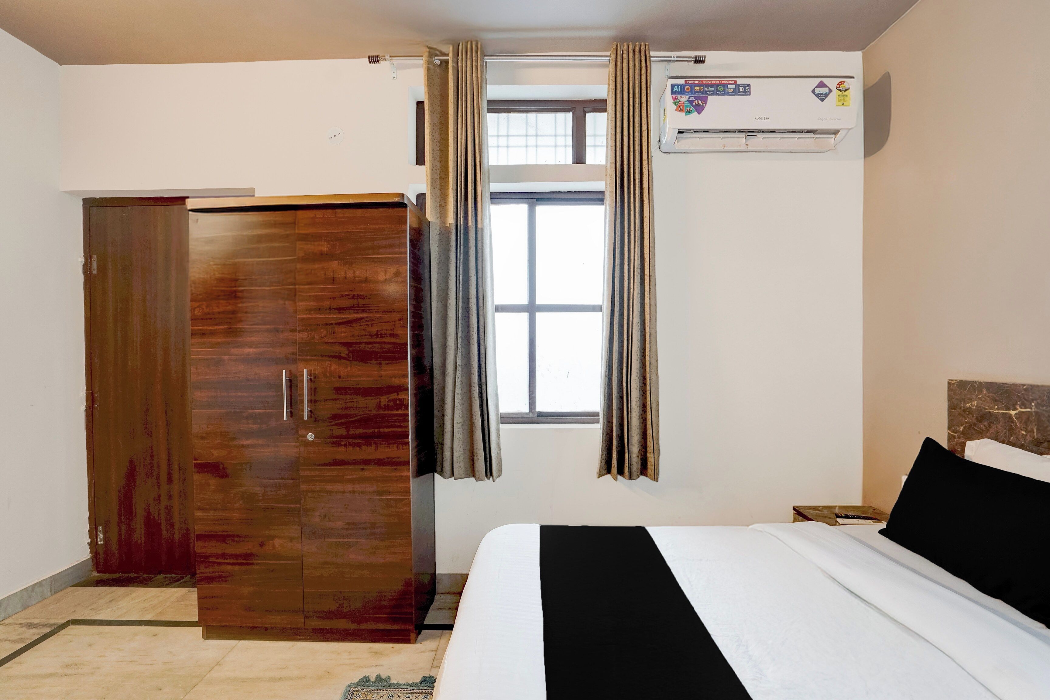 Un dormitorio con un armario de madera, una cama con ropa de cama blanca y negra, un ventanal con cortinas y un equipo de aire acondicionado en la pared.