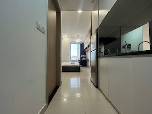 Un pasillo moderno de hotel con una cama, una mesita con flores y una zona de cocina con refrigerador.