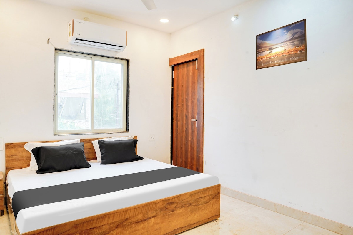 A bedroom with a bed, a window, an air conditioner, a wooden door, and a painting on the wall.