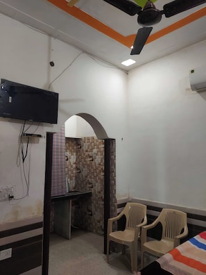 Una habitación con una televisión de pantalla plana montada en la pared, un ventilador de techo y un salpicadero de azulejos. Hay dos sillas de plástico y una cama con colcha a rayas.