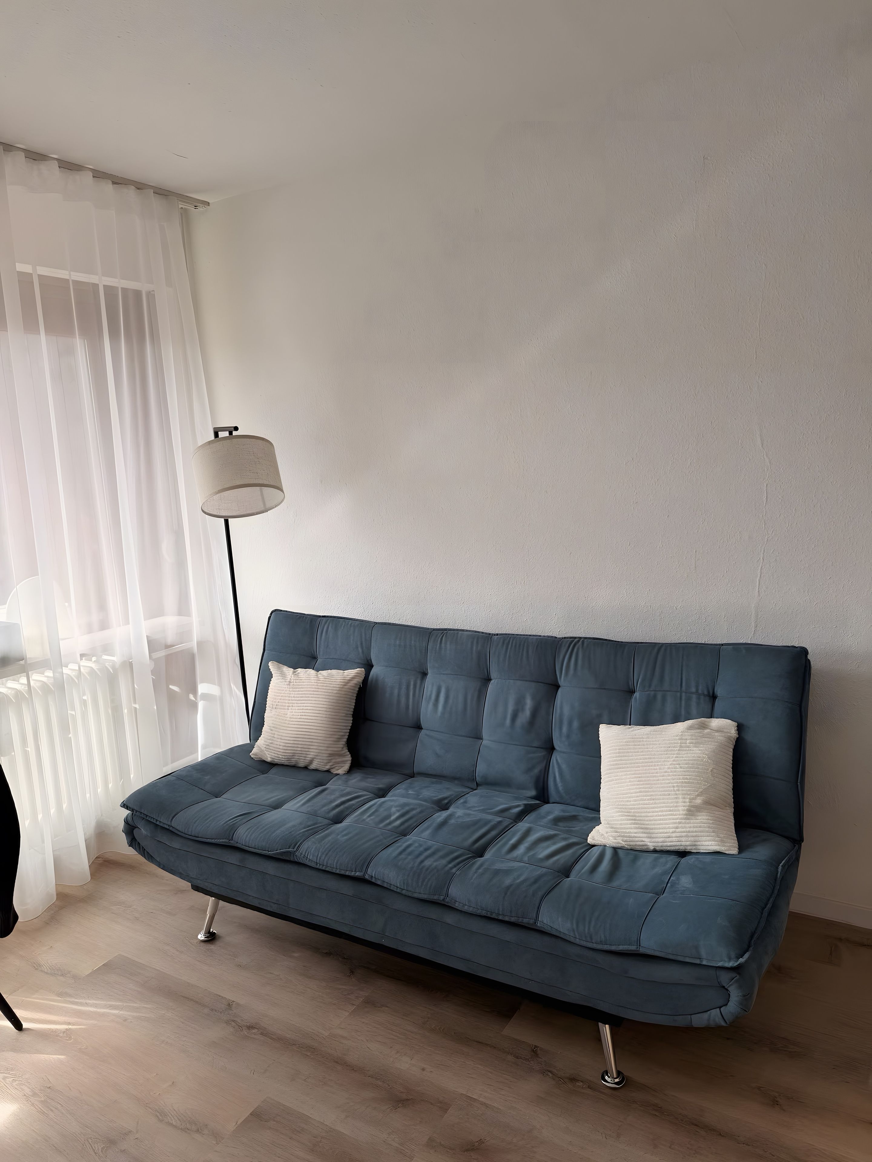 A modern living room with a blue sofa, two white cushions, a floor lamp, and sheer curtains.