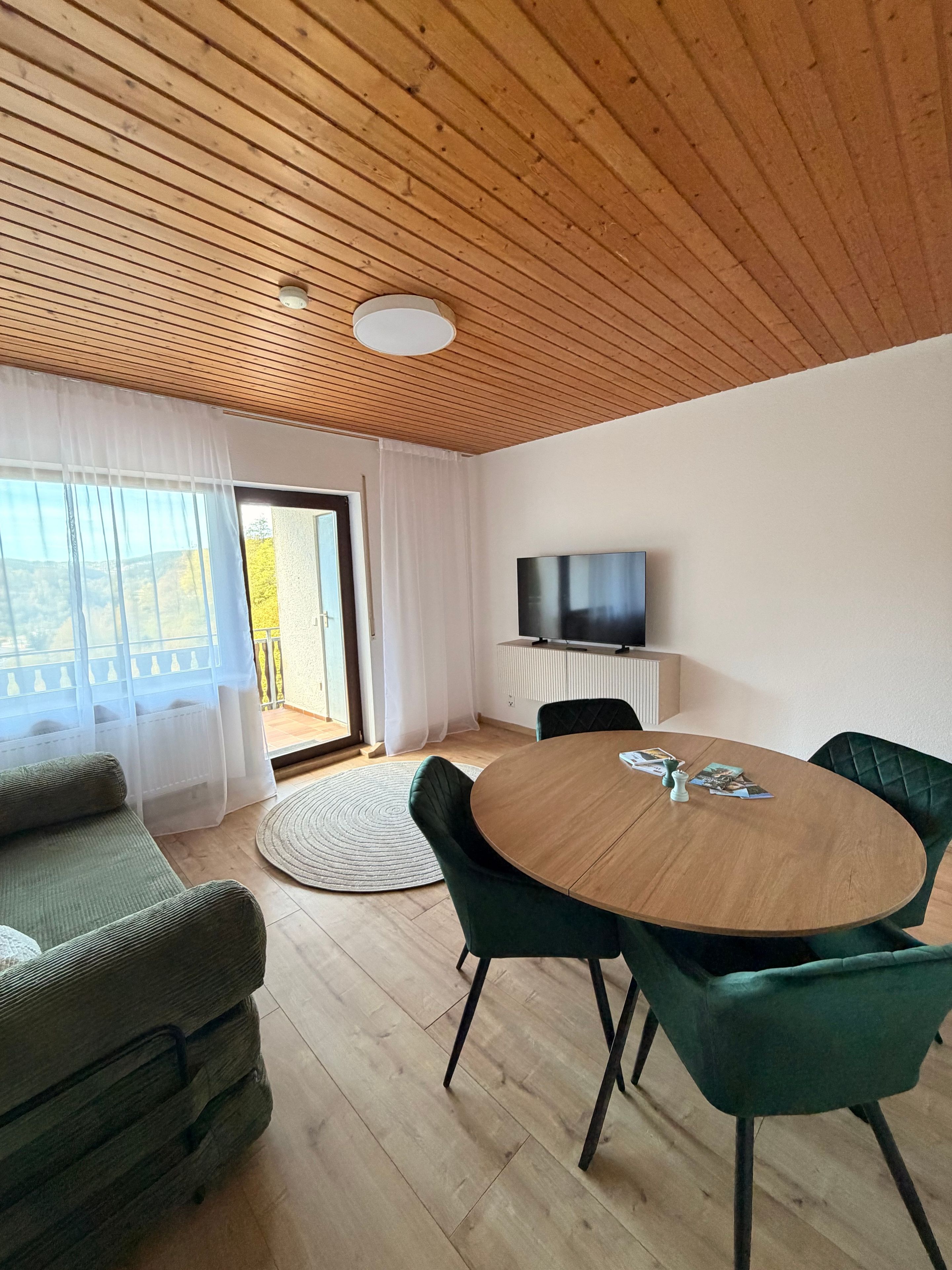 A modern living room with a wooden ceiling, a round table with chairs, a sofa, a television, and a view of the outdoors.