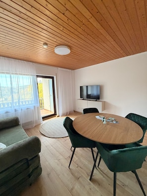 A modern living room with a wooden ceiling, a round table with chairs, a sofa, a television, and a view of the outdoors. - Modern 2-Room Holiday Apartments Triberg (Triberg im Schwarzwald)