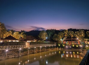 A nighttime resort with a bridge over a calm body of water, palm trees, and illuminated buildings.