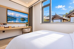 Une chambre d’hôtel comprenant un lit, un bureau avec un ordinateur, une fenêtre donnant sur des bâtiments traditionnels et un tableau représentant un paysage de montagne.