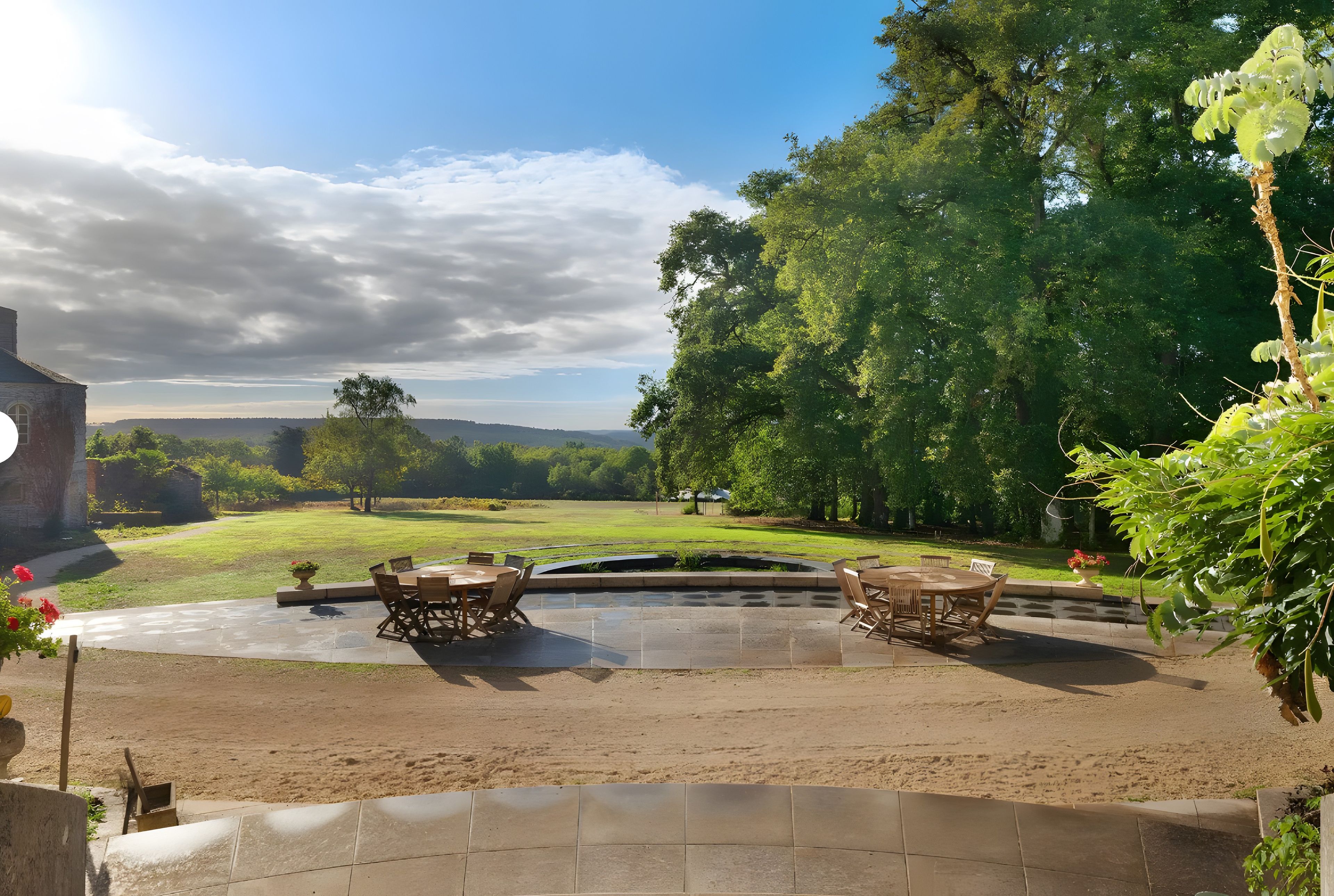 An outdoor patio with two wooden tables and chairs, surrounded by a garden with a view of a distant landscape.