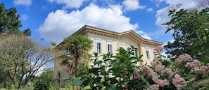 Un edifici gran i antic amb una façana simètrica, envoltat de vegetació exuberanta i plantes florentes.