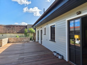 A covered patio area with a wooden deck, a brick wall, and a building with a white exterior and large sliding glass doors. - Holiday Home 'Haus Hovilin' with Private Terrace, Private Garden and Wi-Fi (Höflein bei Bruck an der Leitha)