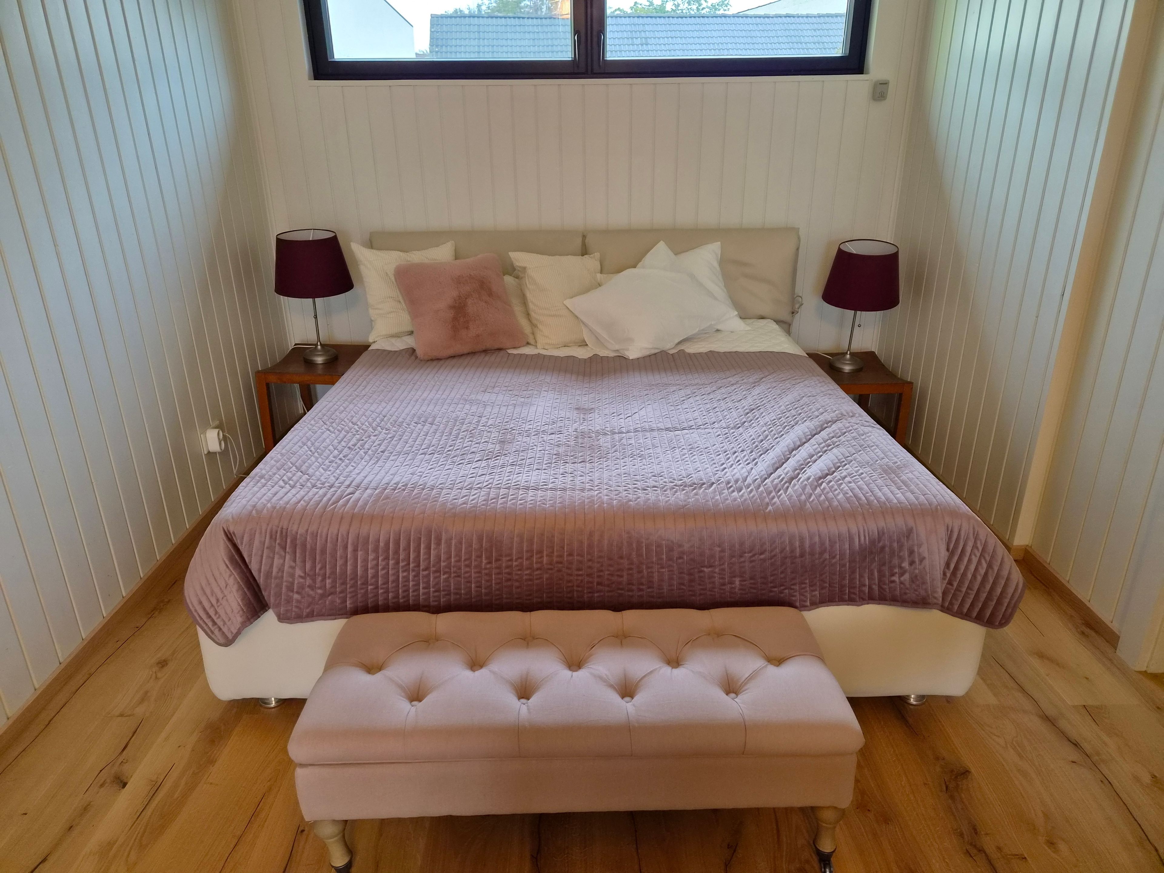 A bedroom with a bed, bedside lamps, a bench, and a window.