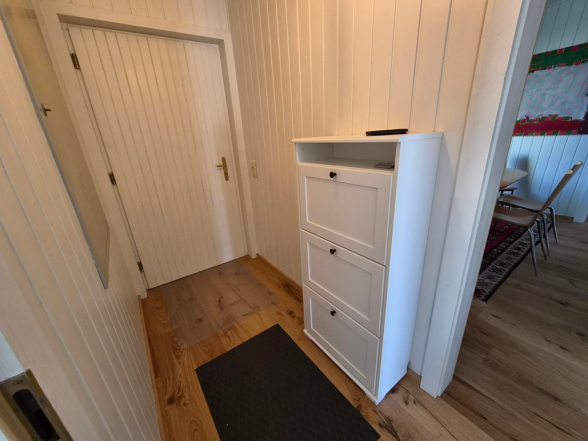 Ein schmaler Flur mit einer weißen Tür, einem weißen Schubladenschrank und einer schwarzen Fußmatte auf dem Holzboden.