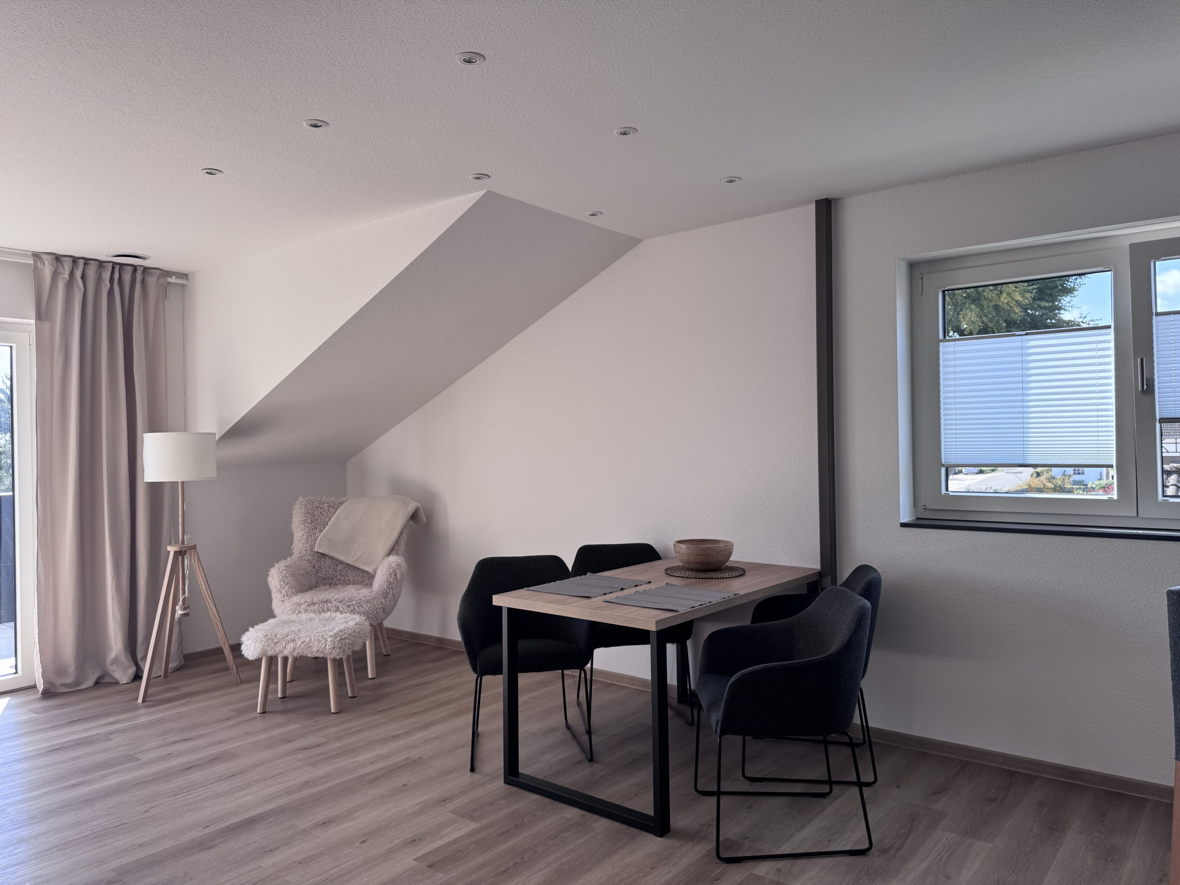 Un espace repas moderne avec une table et des chaises, une lampe et une vue sur l’extérieur par une fenêtre.