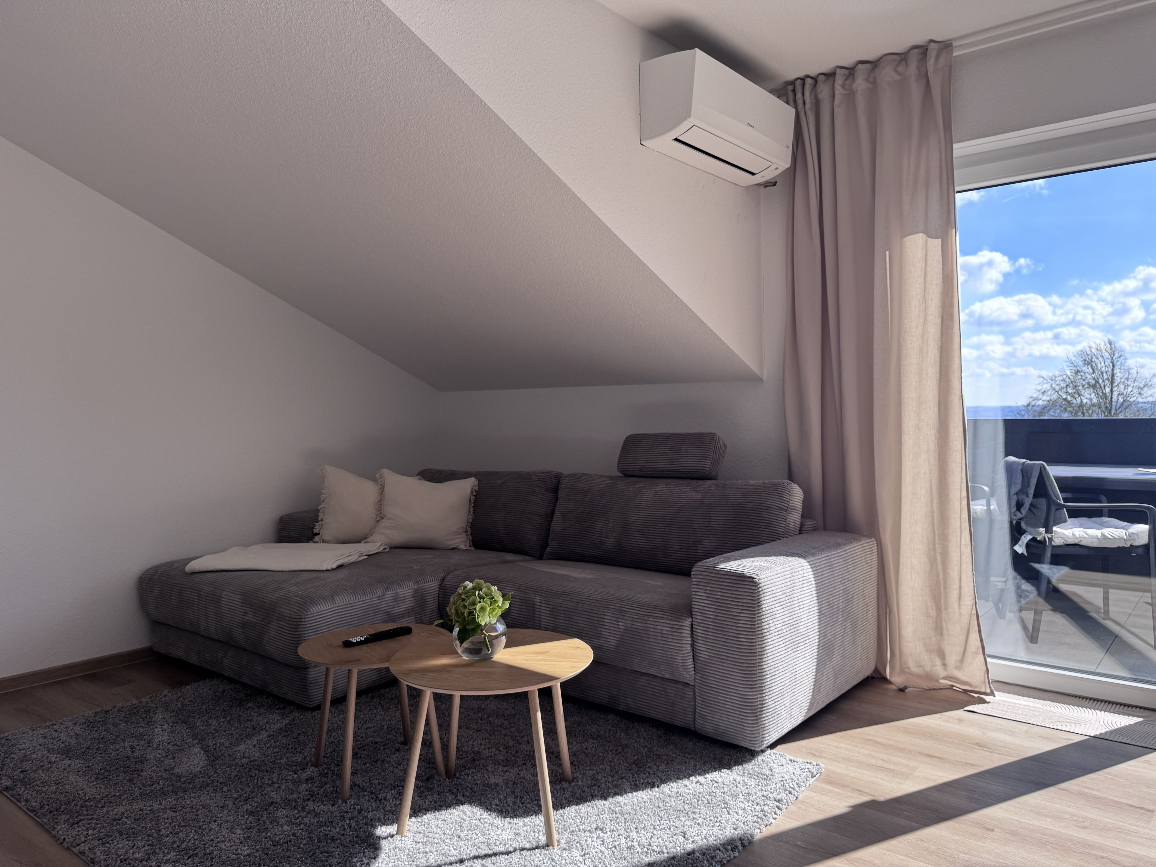 Ein modernes Wohnzimmer mit einer Couch, einem kleinen Tisch mit einer Pflanze und einem großen Fenster mit Vorhängen.