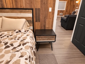 A bedroom with a bed, bedside table, and a view into another room with a couch.